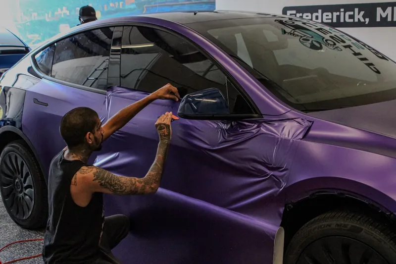 A SpaceShip Wraps installer kneeling beside a Tesla Model Y, working a sheet of purple satin film onto the rear door and quarter panel by hand