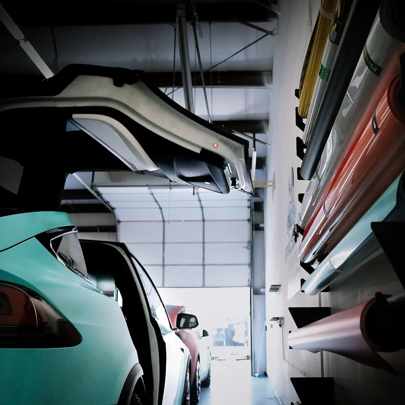 Wide view of the SpaceShip Wraps shop interior, vinyl rolls racked on the wall above a teal wrapped Tesla, roll-up door open at the end of the bay